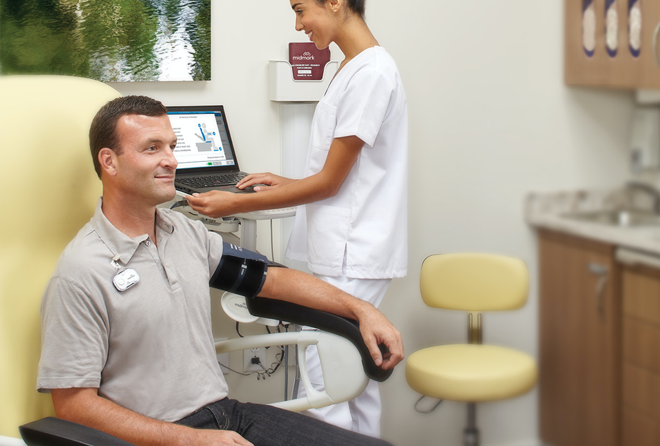 A nurse on a laptop beside a patient in an exam chair with a blood pressure cuff.