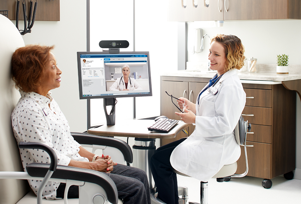 A doctor and patient in an exam room. The doctor is consulting with another doctor virtually on a video call on a computer from the workstation