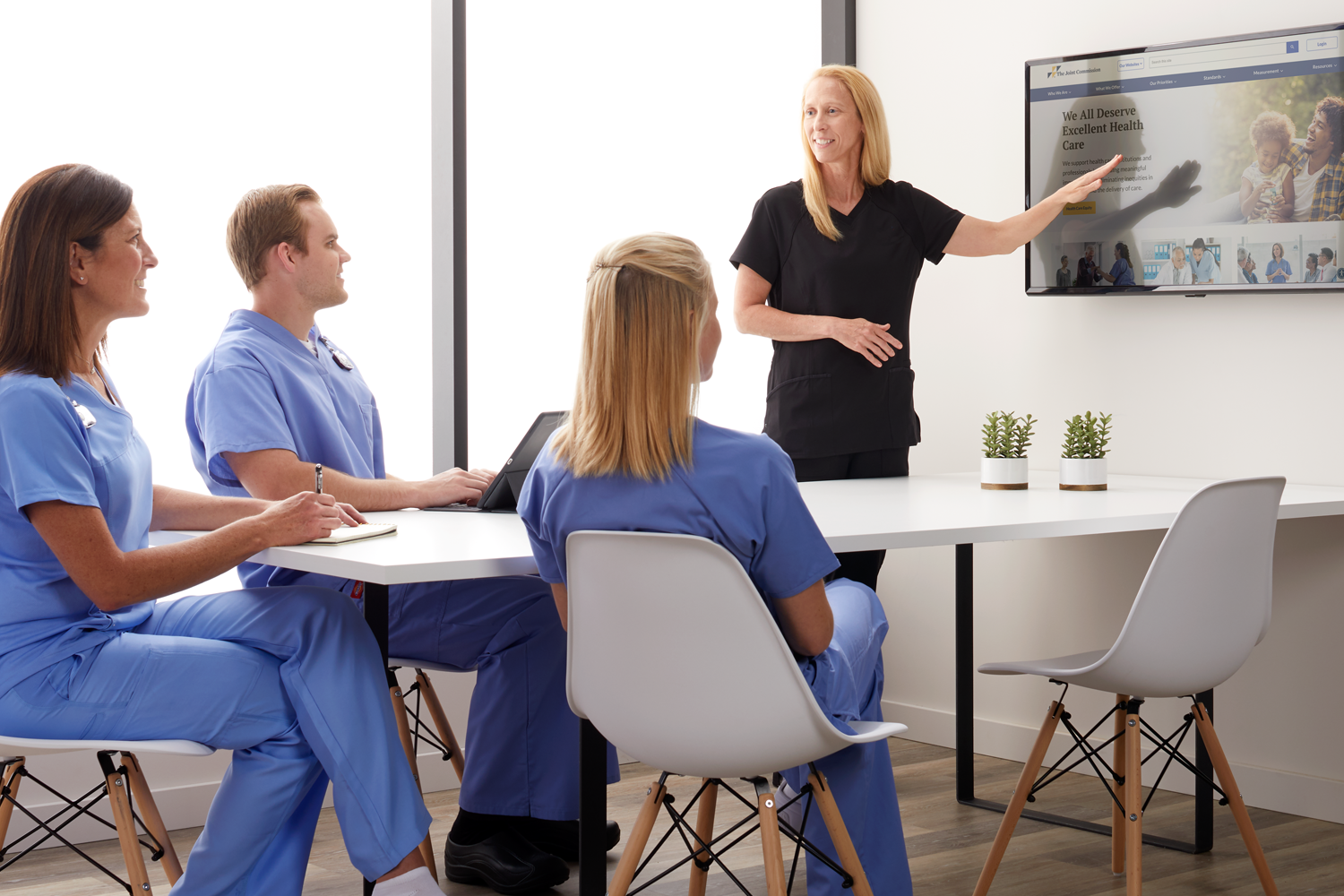 Multiple nurses sat at a table looking at an instructor, pointing at a screen